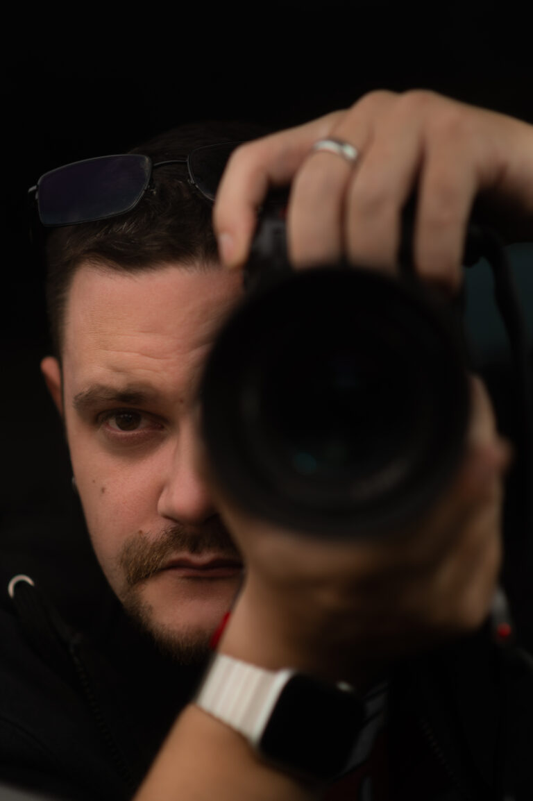 Osoba v tmavé bundě a náramkových hodinkách drží fotoaparát u obličeje a soustředěně zaostřuje. Na hlavě mají brýle a na levé ruce nosí prsten. Obraz je slabě osvětlen s důrazem na oči osoby a čočku fotoaparátu.