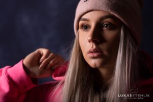 Člověk se zamyšleně dívá do strany, má na sobě světle růžovou čepici a jasně růžovou mikinu s kapucí. Rukou se lehce dotýkají kapoty. Pozadí je tmavé a rozmazané, což zvyšuje zaměření na jejich klidný výraz a dlouhé světlé vlasy.