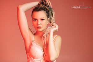Žena se spletenými vlasy pózuje na korálovém pozadí a má na sobě růžový krajkový top. Levou paži má zvednutou a kruhové náušnice jí dodávají na vzhledu. Osvětlení vrhá teplou, měkkou záři a vytváří stylovou a sebevědomou atmosféru. Fotografoval Lukáš Stehlík.