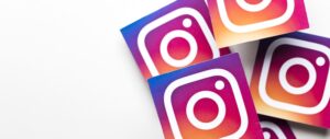 Na bílém povrchu je roztroušeno několik nálepek s logem Instagramu s přechodem růžové, oranžové a žluté. Loga jsou čtvercová se zaobleným designem fotoaparátu v bílé barvě.