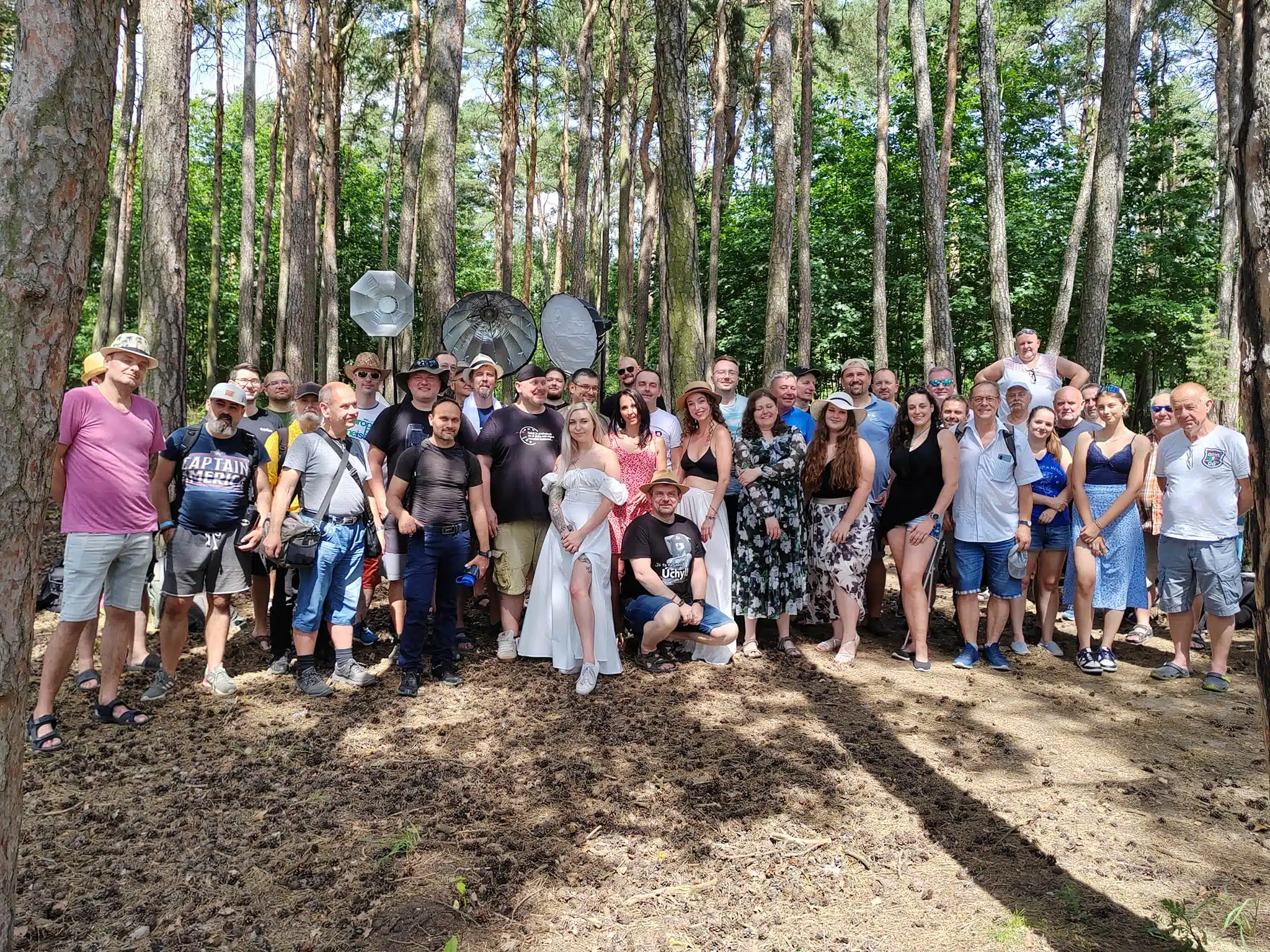 Velká skupina lidí pózuje pro fotografii v zalesněné oblasti s osvětlovacím zařízením viditelným v pozadí za slunečného dne.