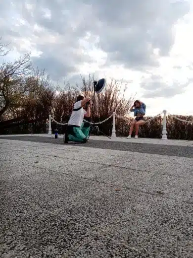 lukasstehlik-00004 Fotograf klečí na zemi a fotí sedící ženu venku, v pozadí zatažená obloha a keře.