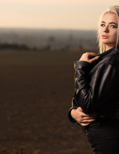 Žena s dlouhými blond vlasy v černé kožené bundě stojí na otevřeném poli při západu slunce a dívá se do strany.