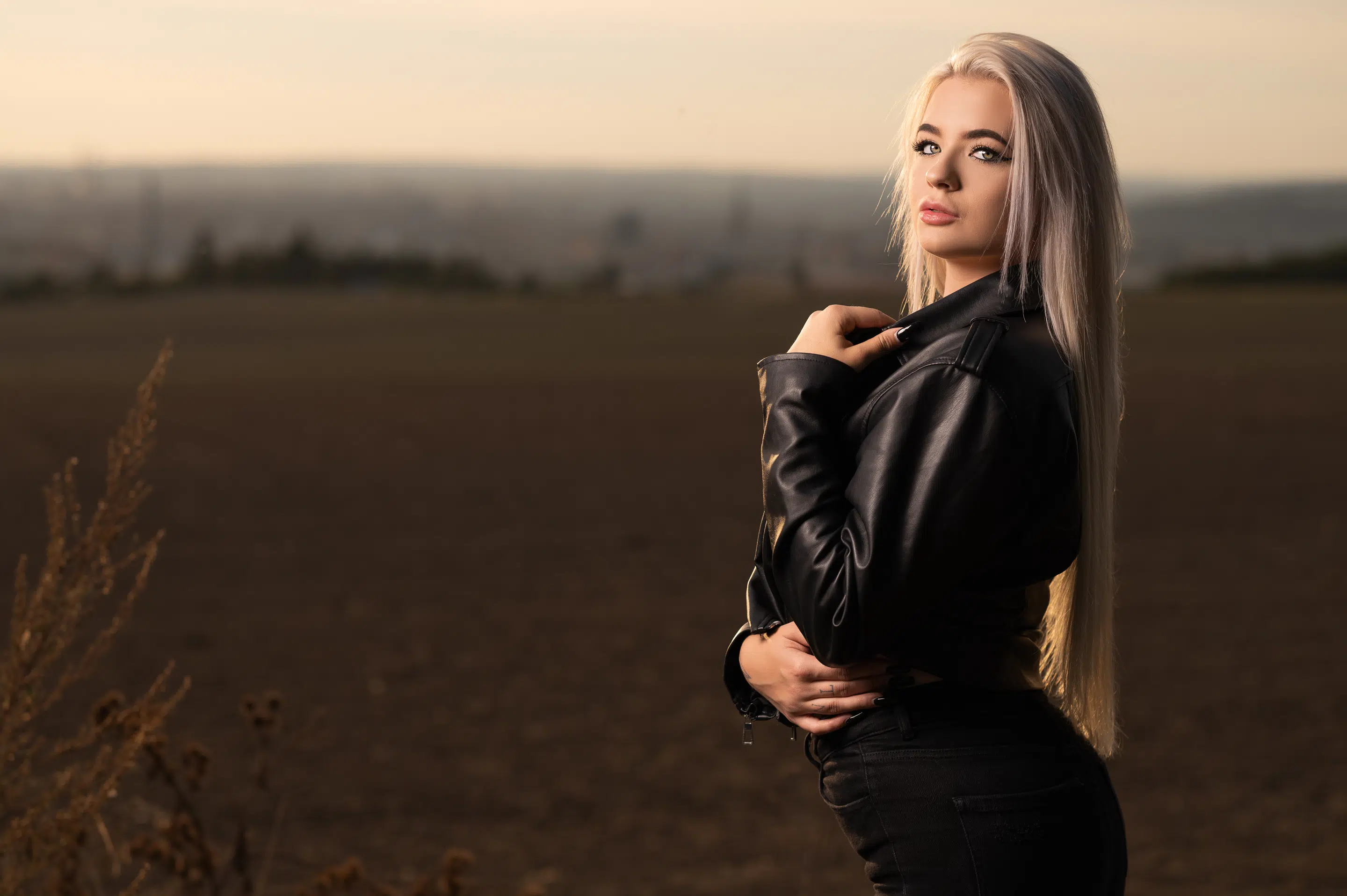 Žena s dlouhými blond vlasy v černé kožené bundě stojí na otevřeném poli při západu slunce a dívá se do strany.