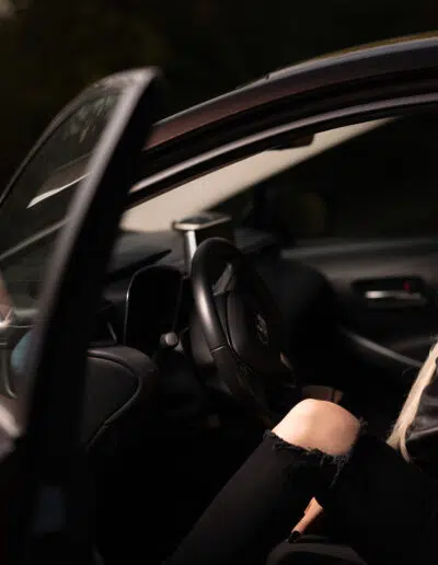 Žena s dlouhými blond vlasy sedí na sedadle řidiče v autě s otevřenými dveřmi, má na sobě černou koženou bundu a roztrhané černé džíny a vypadá uvolněně.