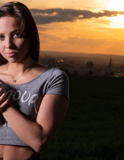 Žena s hnědými vlasy stojí na poli při západu slunce, má na sobě šedý crop top a náhrdelník, ruce má sepjaté u hrudi.