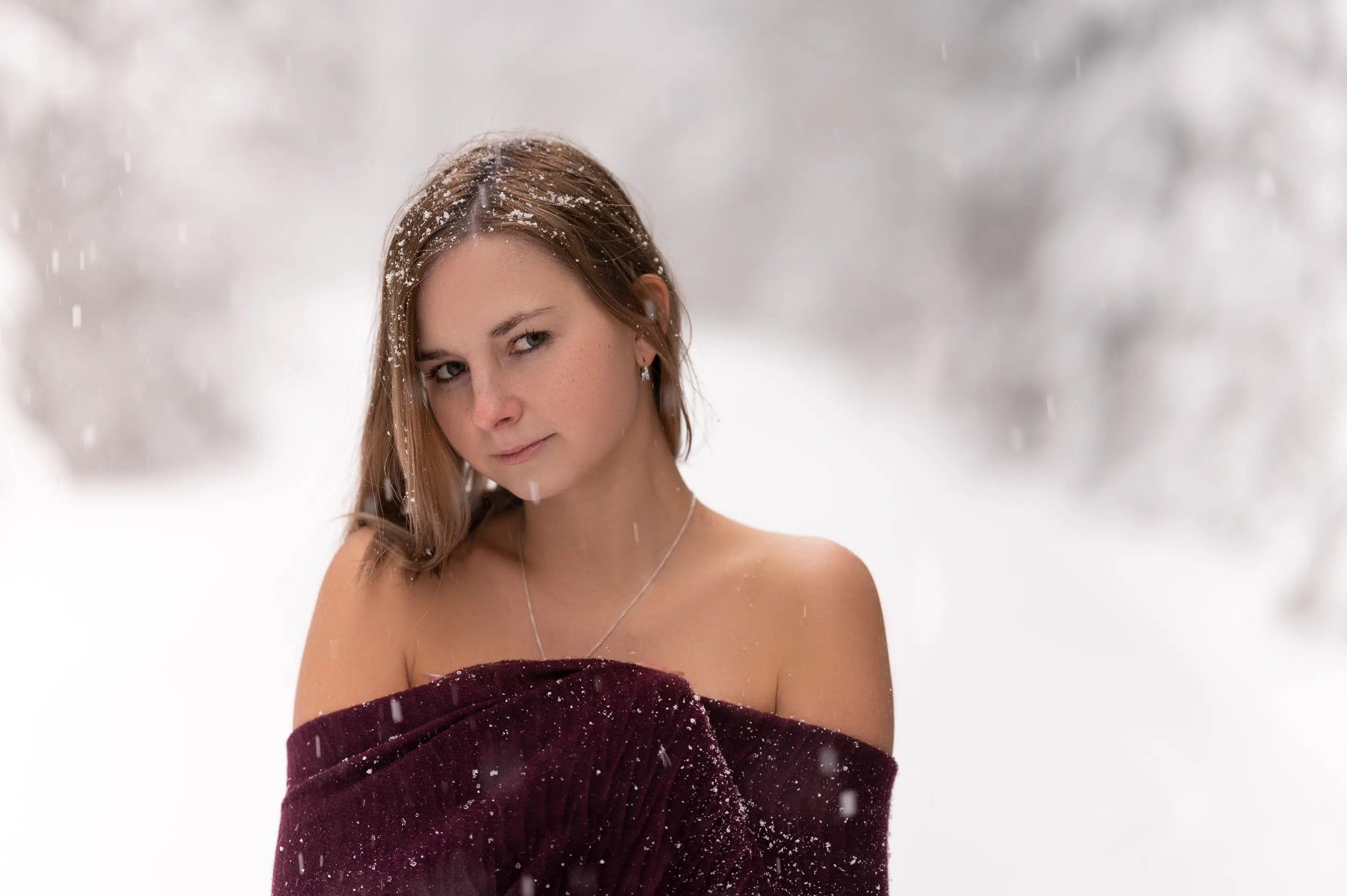 lukasstehlik-gallery-00007 Žena stojí venku v padajícím sněhu, má na sobě tmavý top s odhalenými rameny. Sněhové vločky jí leží na vlasech a ramenou; pozadí je rozmazané stromy pokrytými sněhem.