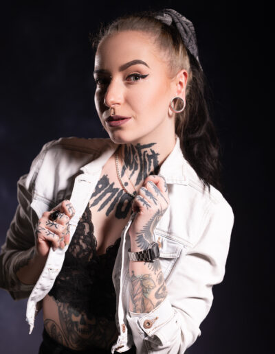 Žena s tetováním a piercingy pózuje na tmavém pozadí, má na sobě bílou džínovou bundu přes černý krajkový top.