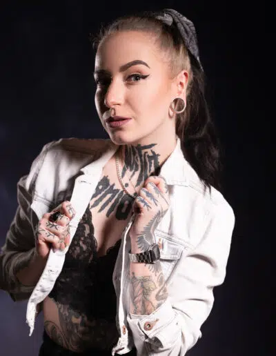 Žena s tetováním a piercingy pózuje na tmavém pozadí, má na sobě bílou džínovou bundu přes černý krajkový top.