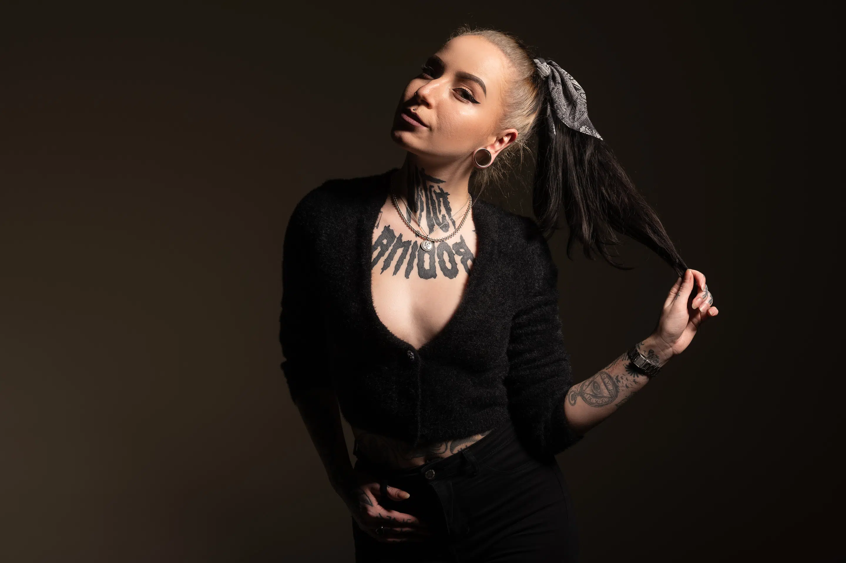 lukasstehlik-gallery-00010 Žena s tetováním a piercingy v černém svetru a tmavých kalhotách si drží dlouhý culík a pózuje na tmavém pozadí.