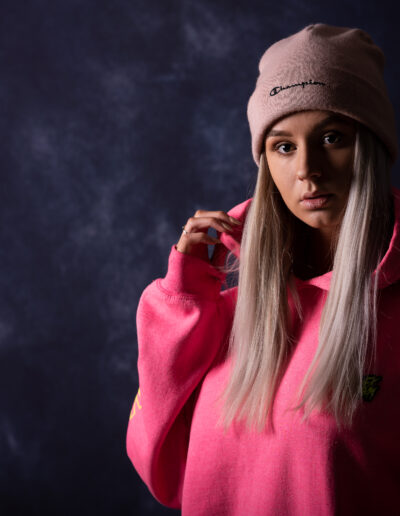 Osoba s dlouhými blond vlasy má na sobě růžovou mikinu s kapucí a světle růžovou čepici a stojí na tmavém, texturovaném pozadí.