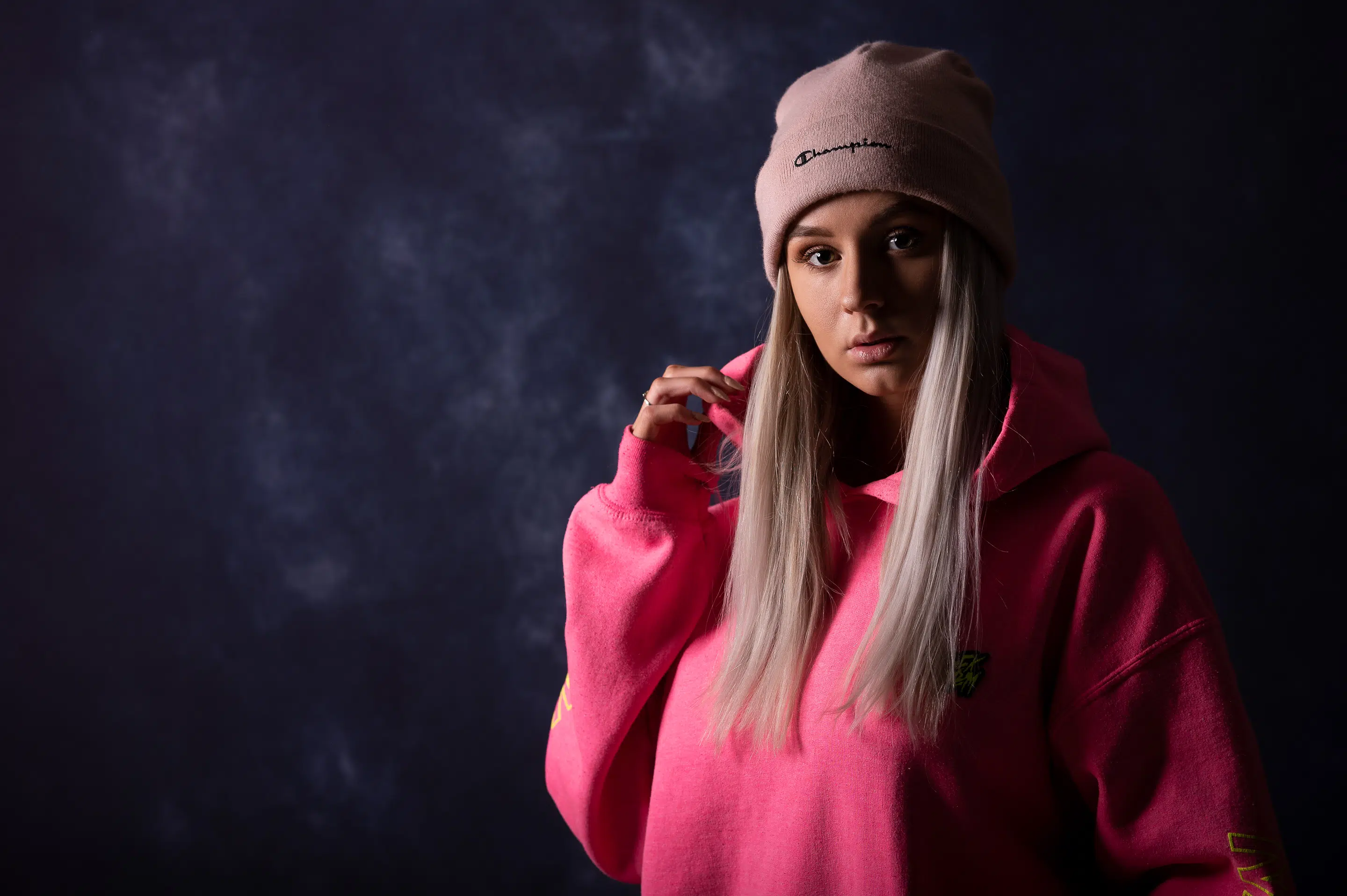 lukasstehlik-gallery-00014 Osoba s dlouhými blond vlasy má na sobě růžovou mikinu s kapucí a světle růžovou čepici a stojí na tmavém, texturovaném pozadí.