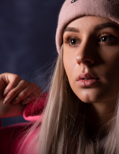 Žena s dlouhými blond vlasy má na sobě růžovou čepici a růžovou mikinu s kapucí a na tmavém pozadí se dívá mírně stranou.