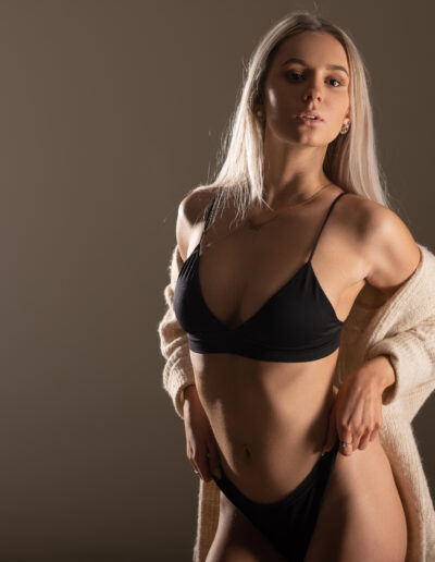 Žena s dlouhými blond vlasy pózuje v černých bikinách a béžovém svetru na prostém, jemně osvětleném pozadí.