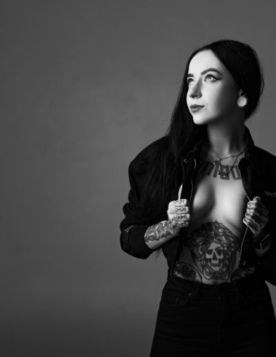 Žena s tetováním pózuje v částečně rozepnuté bundě a dívá se do strany na prostém černobílém pozadí.