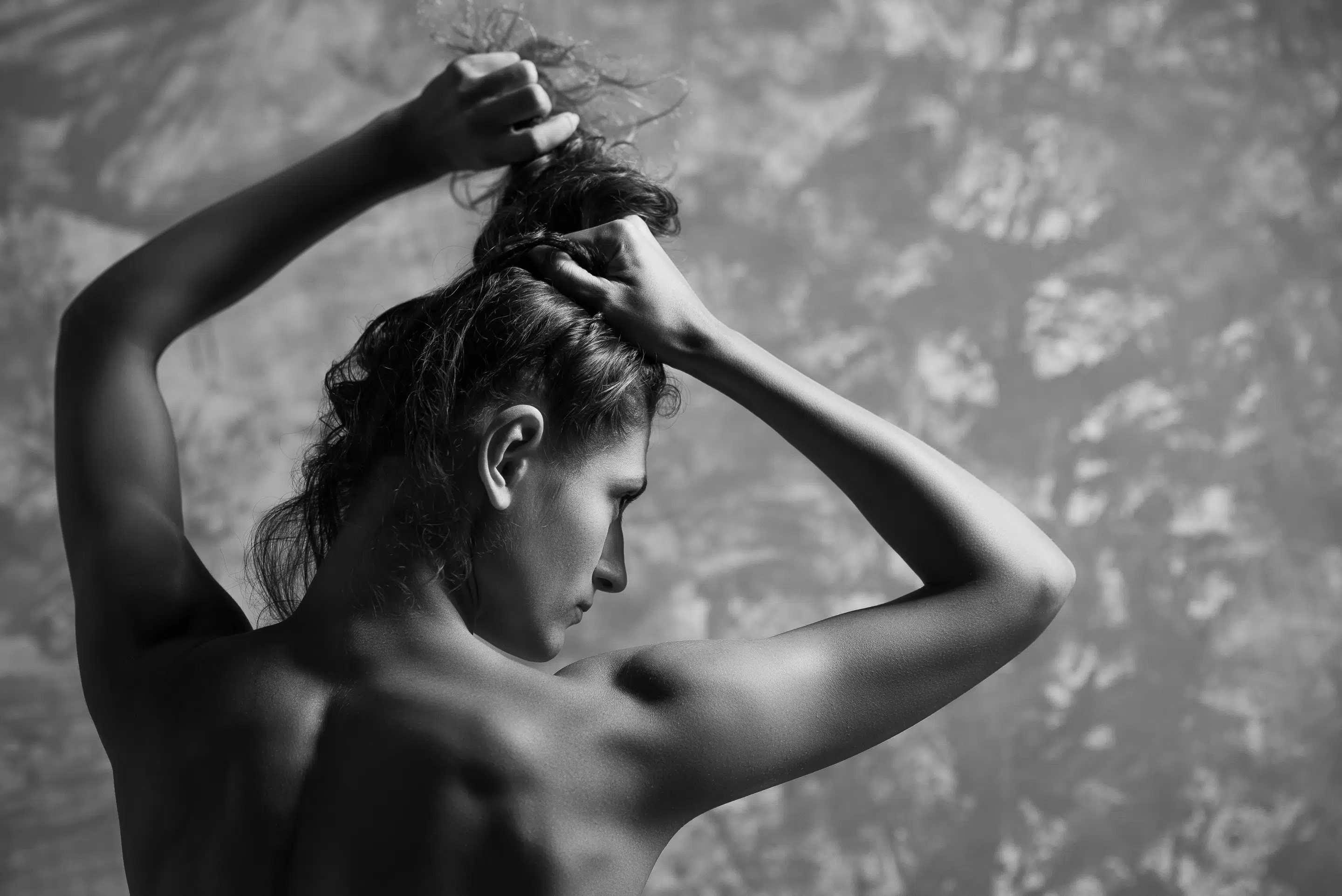 lukasstehlik-gallery-00034 Černobílá fotografie osoby s odhalenými rameny odvrácenými, zdviženýma rukama, která si svazuje vlasy na texturovaném pozadí.