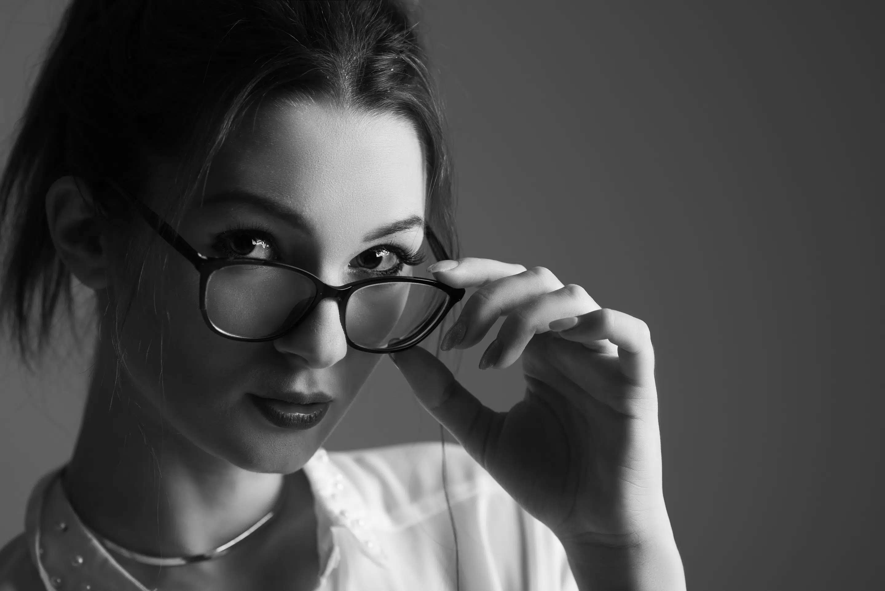 Černobílá fotografie ženy s tmavými vlasy, která se dívá do fotoaparátu, v jedné ruce si drží brýle a má na sobě košili s límečkem a tenký náhrdelník.