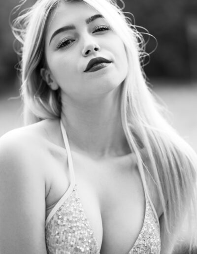 Žena s dlouhými blond vlasy v flitrovaném topu pózuje venku a dívá se přímo do kamery. Fotografie je černobílá.
