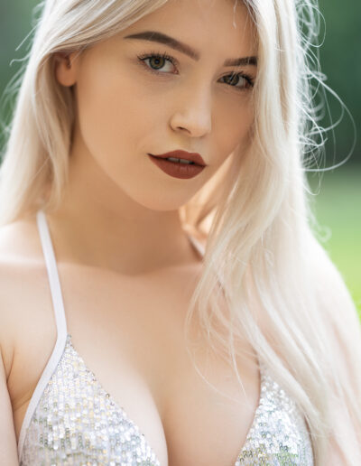 Žena s dlouhými blond vlasy v flitrovaném bikinovém topu stojí venku se zelení v rozmazaném pozadí.