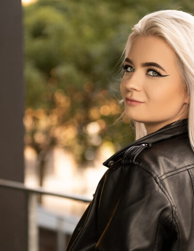 Žena s dlouhými blond vlasy a černou koženou bundou stojí venku a dívá se přes rameno, zatímco v rozmazaném pozadí jsou vidět stromy.