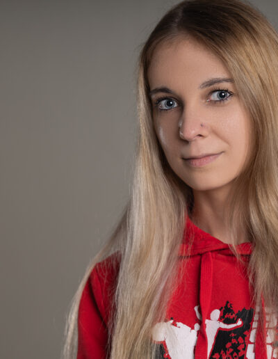 Žena s dlouhými blond vlasy má na sobě červenou mikinu s bíločernou grafikou a stojí na prostém šedém pozadí.