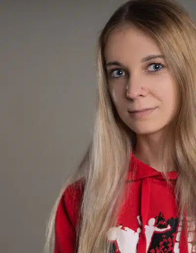 Žena s dlouhými blond vlasy má na sobě červenou mikinu s bíločernou grafikou a stojí na prostém šedém pozadí.