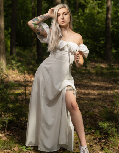 Žena s dlouhými blond vlasy a tetováním pózuje v bílých šatech s odhalenými rameny a teniskách ve sluncem zalitém lese.