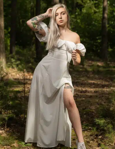 Žena s dlouhými blond vlasy a tetováním pózuje v bílých šatech s odhalenými rameny a teniskách ve sluncem zalitém lese.