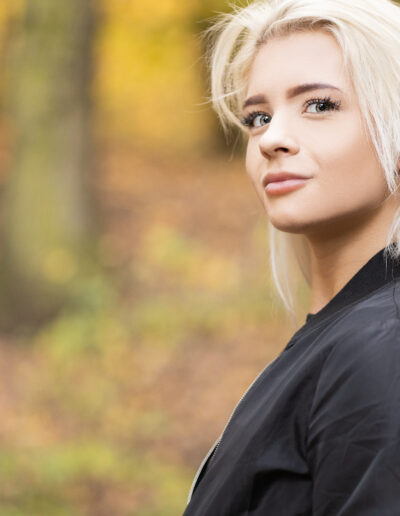 Žena s dlouhými blond vlasy v černé bundě stojí venku v lese s rozmazaným podzimním listím v pozadí.