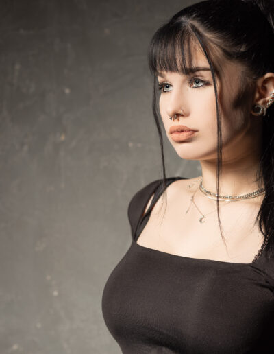 Žena s dlouhými černými vlasy sepnutými do culíku, v černém topu, se stříbrnými náhrdelníky a piercingy, stojí na šedém texturovaném pozadí.