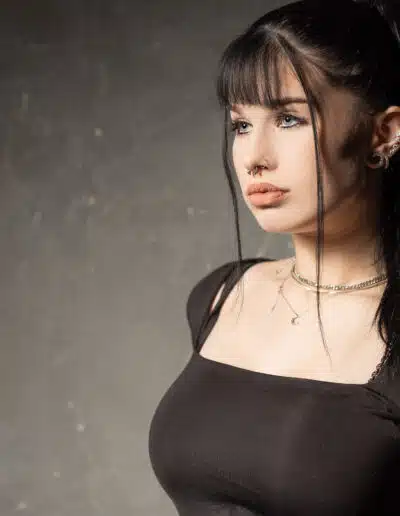 Žena s dlouhými černými vlasy sepnutými do culíku, v černém topu, se stříbrnými náhrdelníky a piercingy, stojí na šedém texturovaném pozadí.