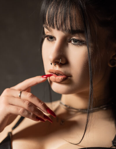 Žena s tmavými vlasy a ofinou, která má na sobě septum prsten, se prstem dotýká rtů. Má dlouhé červené nehty a je oblečená v černém topu s viditelnými šperky.